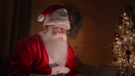 Der-Weihnachtsmann-Arbeitet-Zu-Hause-Am-Computer-Mit-Brille-Und-Bart.-Zauberhafte-Weihnachtslichter-Auf-Dem-Hintergrund-Eines-Geschmückten-Weihnachtsbaums